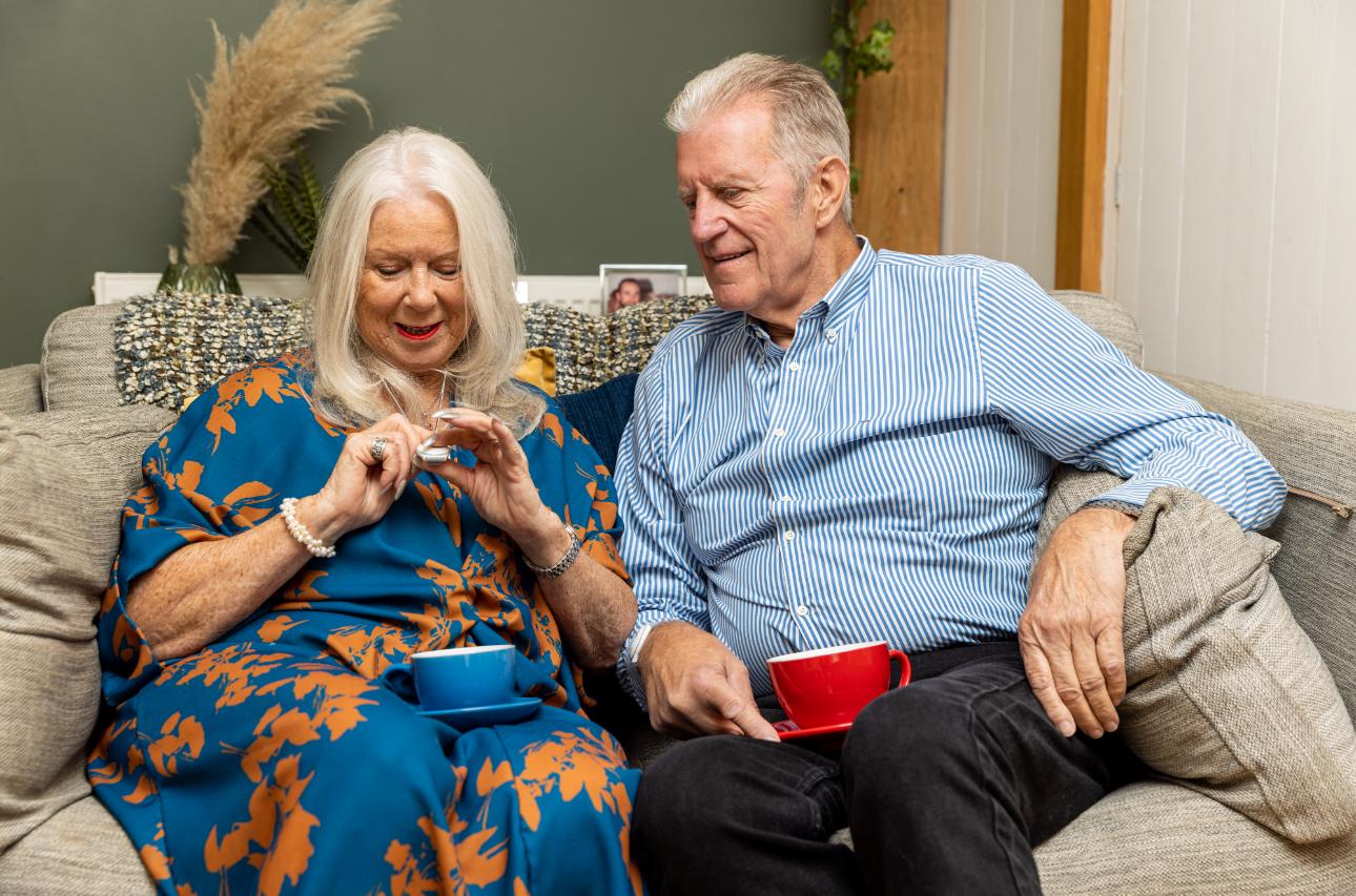 An older couple relaxing together on the sofa, both wearing their Red Button pendants