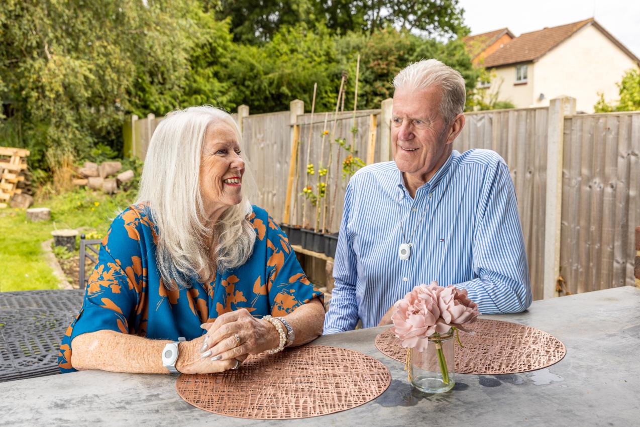 An older couple enjoying time together in their garden, both wearing Red Button personal alarms